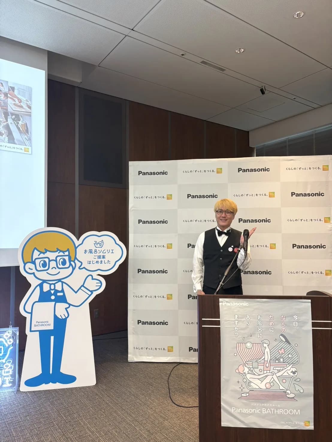 「お風呂ソムリエ」のパネルの横で、演台に立つお風呂のソムリエ 松永武